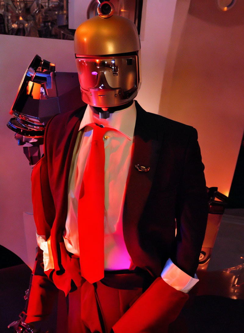 Gold-helmeted humanoid robot in black suit with red tie and visor, arm raised under red