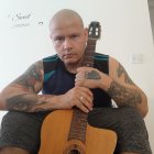 Shaved Head Person Holding Ornate Guitar in Embroidered Jacket