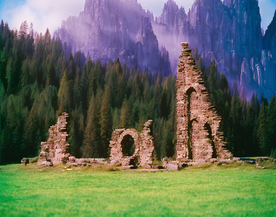 Ruined stone structures in green meadow with forest and mountains.