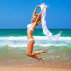 Woman in bikini leaping on sandy beach with sheer scarf, blue skies, turquoise water