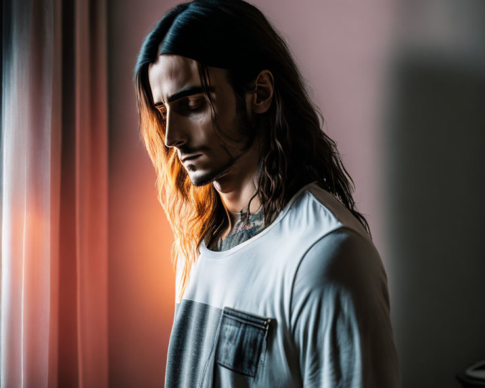 Long-haired bearded man gazes down by window in soft light