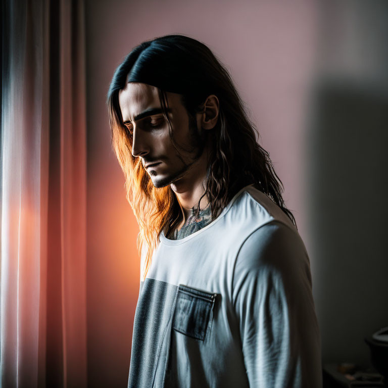 Long-haired bearded man gazes down by window in soft light