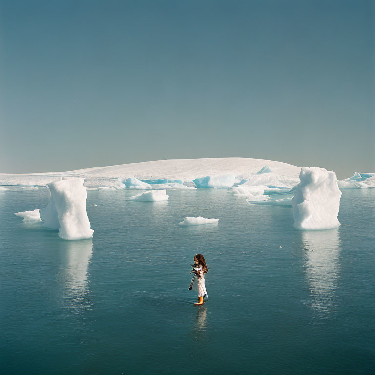 Person standing in tranquil waters surrounded by icebergs under clear blue sky