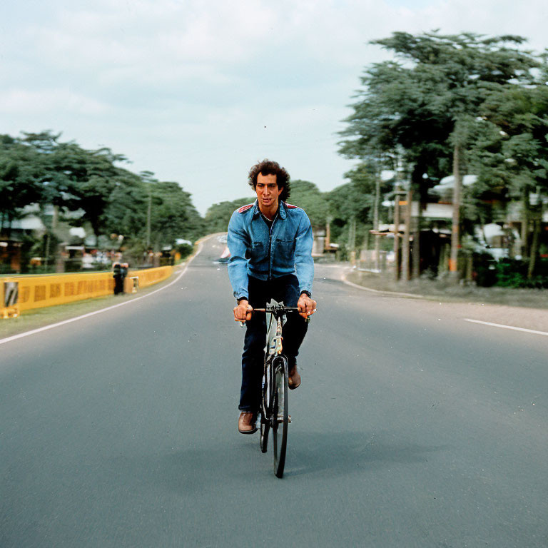 Man in Blue Jacket Riding Bicycle on Empty Road with Trees and Cloudy Sky