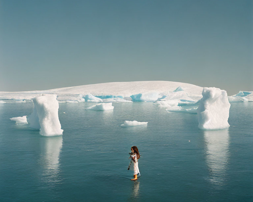 Person standing in tranquil waters surrounded by icebergs under clear blue sky