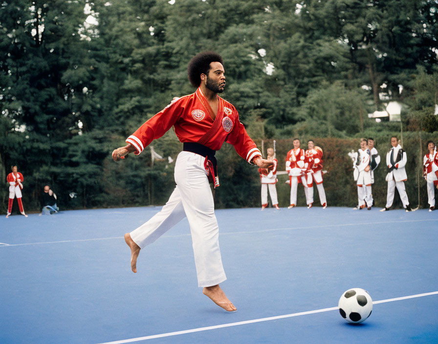 Person in red and black martial arts uniform kicks near soccer ball on blue surface with onlookers in