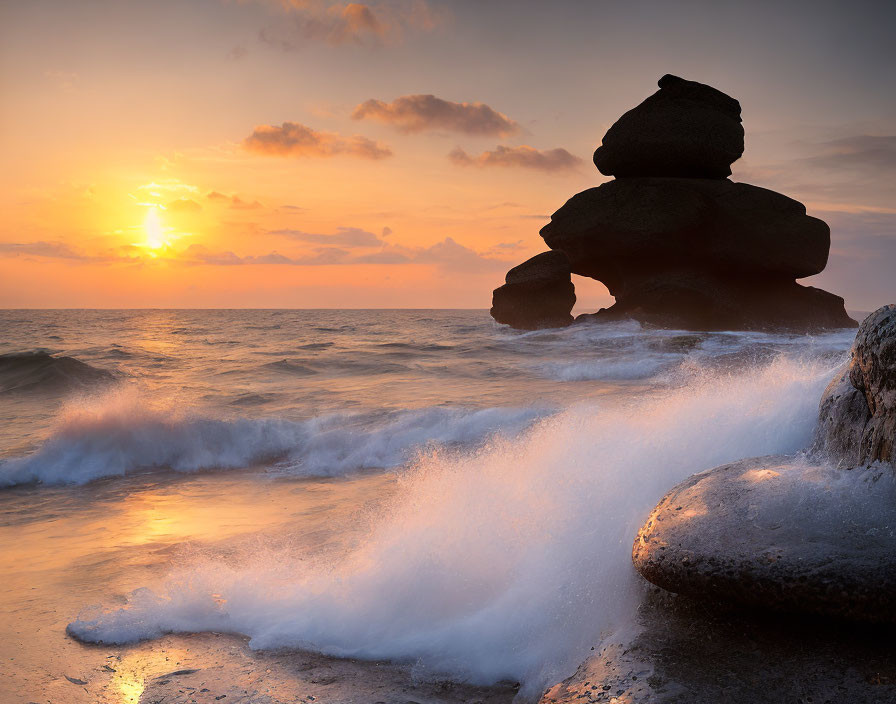 Rocky beach sunset with crashing waves and unique rock formation.