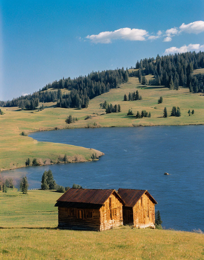 Rustic wooden cabins by serene lake, rolling hills, trees, blue sky