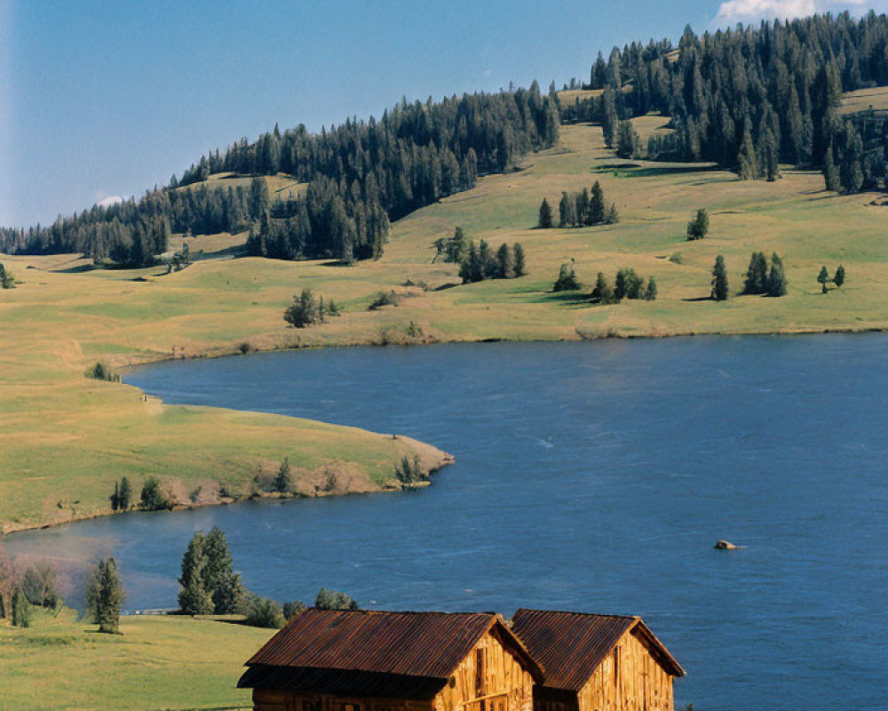 Rustic wooden cabins by serene lake, rolling hills, trees, blue sky