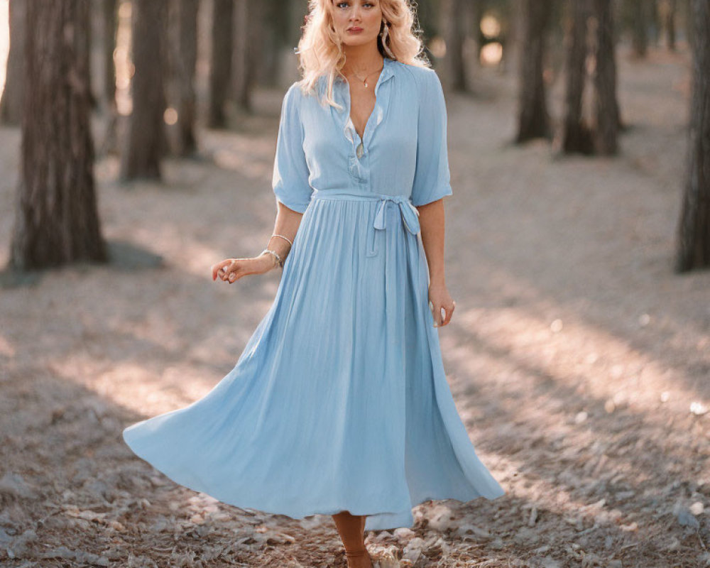 Woman in flowing blue dress in sun-dappled forest with blond hair and simple jewelry.