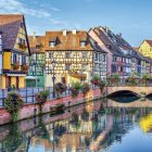 Detailed artwork of quaint houses by a calm river with boats and vibrant foliage.