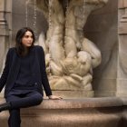 Fashionable woman in trench coat and hat sitting on weathered stairway outside classical building.