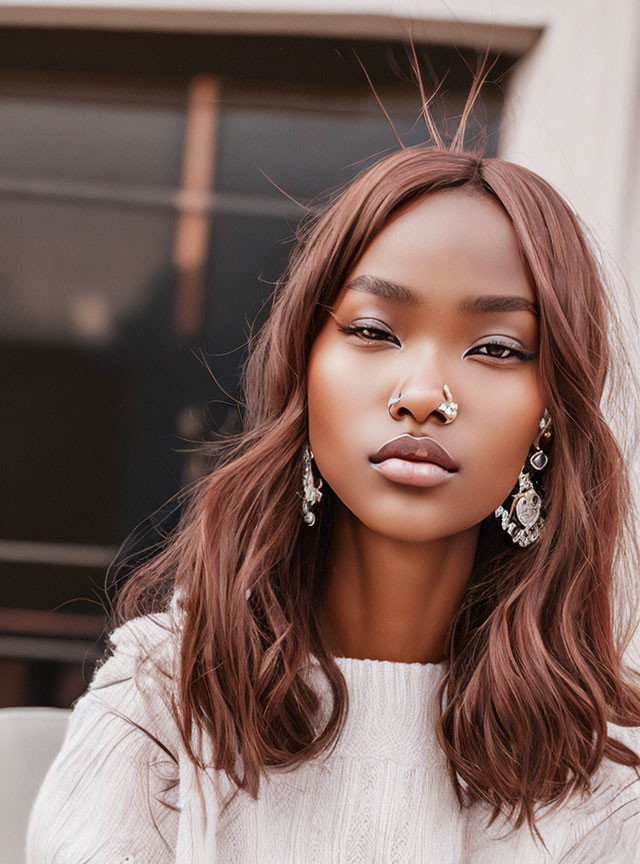 Woman with septum piercing and earrings in white sweater, gazing at camera with loose waves.