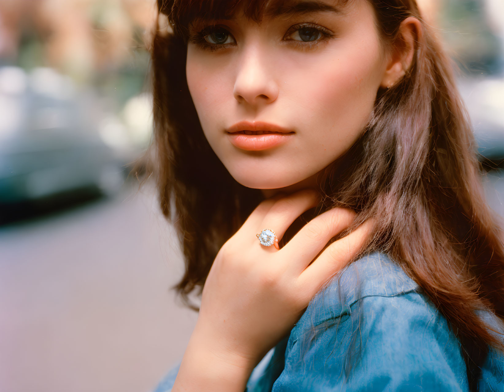 Brunette woman in blue shirt with sparkling ring, blurred cars background