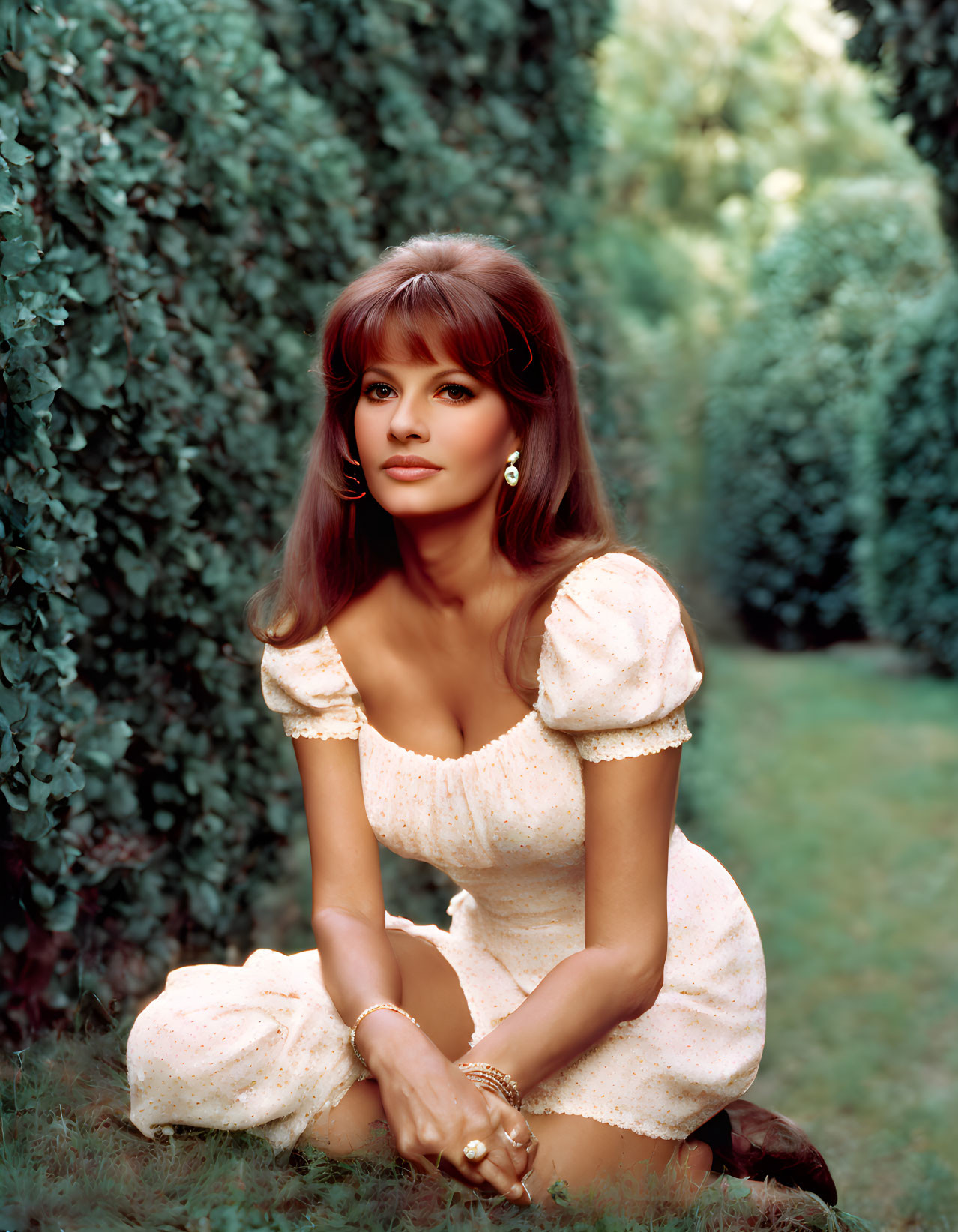 Brunette woman in vintage white dress crouching in lush garden.