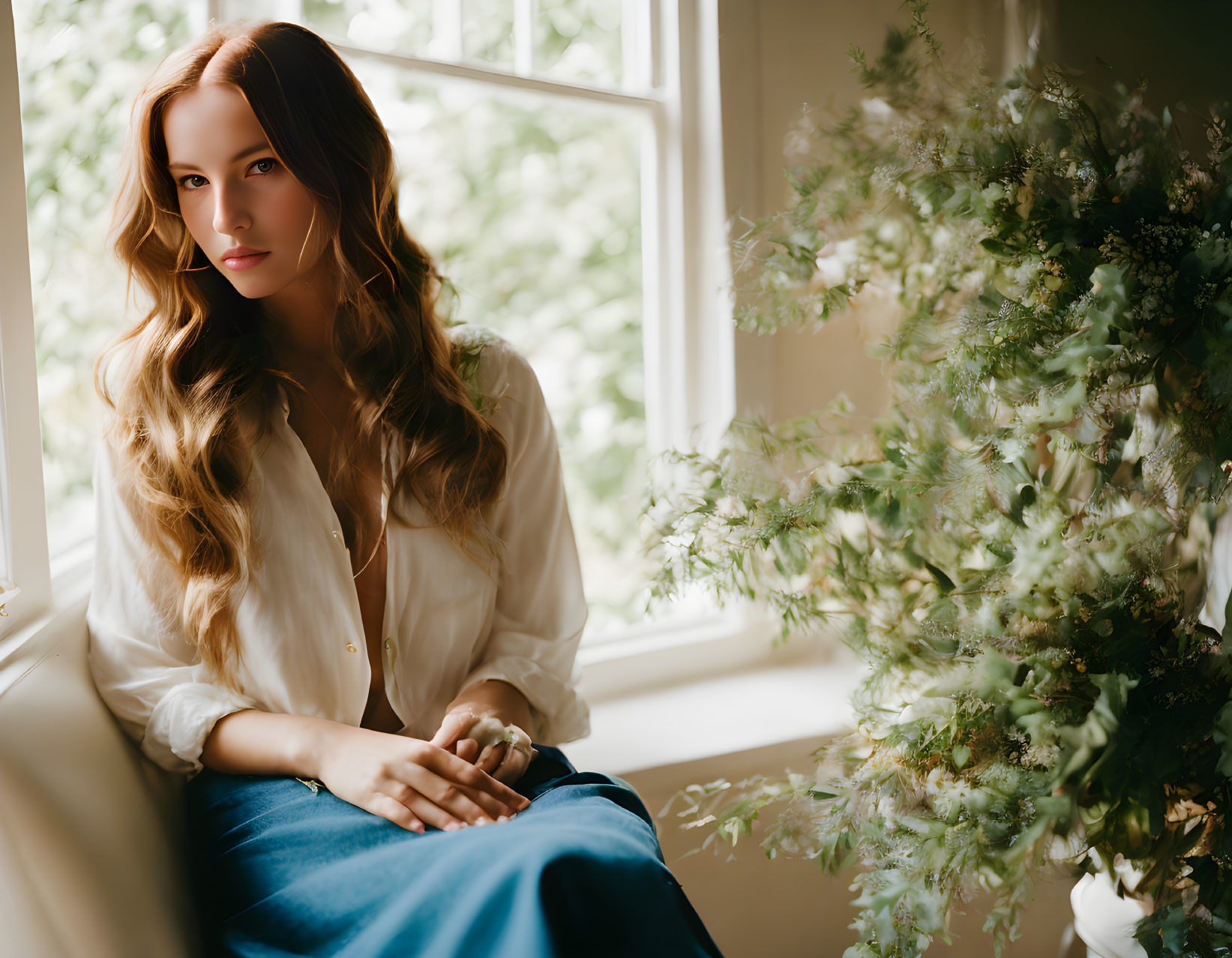 Woman with flowing hair in white shirt and blue skirt sitting by window with green foliage