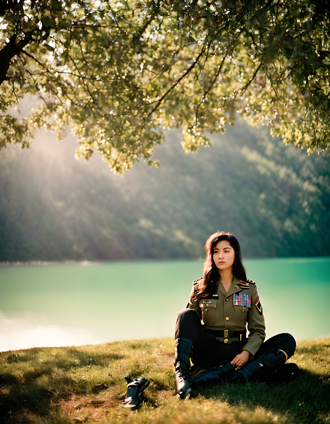 Military person relaxes by lake under tree with sunlight.