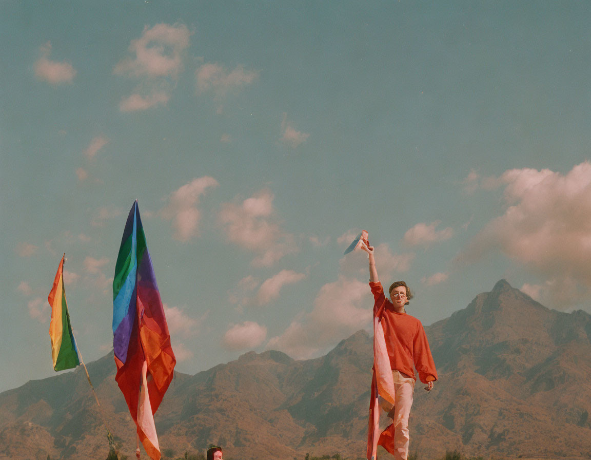 Person in orange fabric with peace sign near colorful flags and mountains.