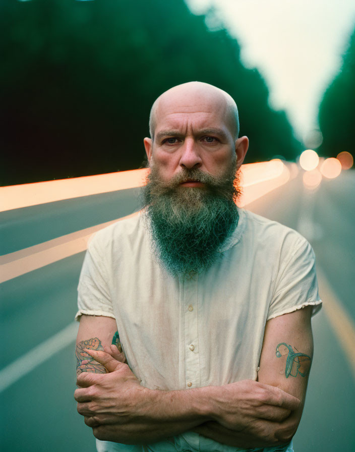 Bald man with gray beard and tattoos standing on road at twilight