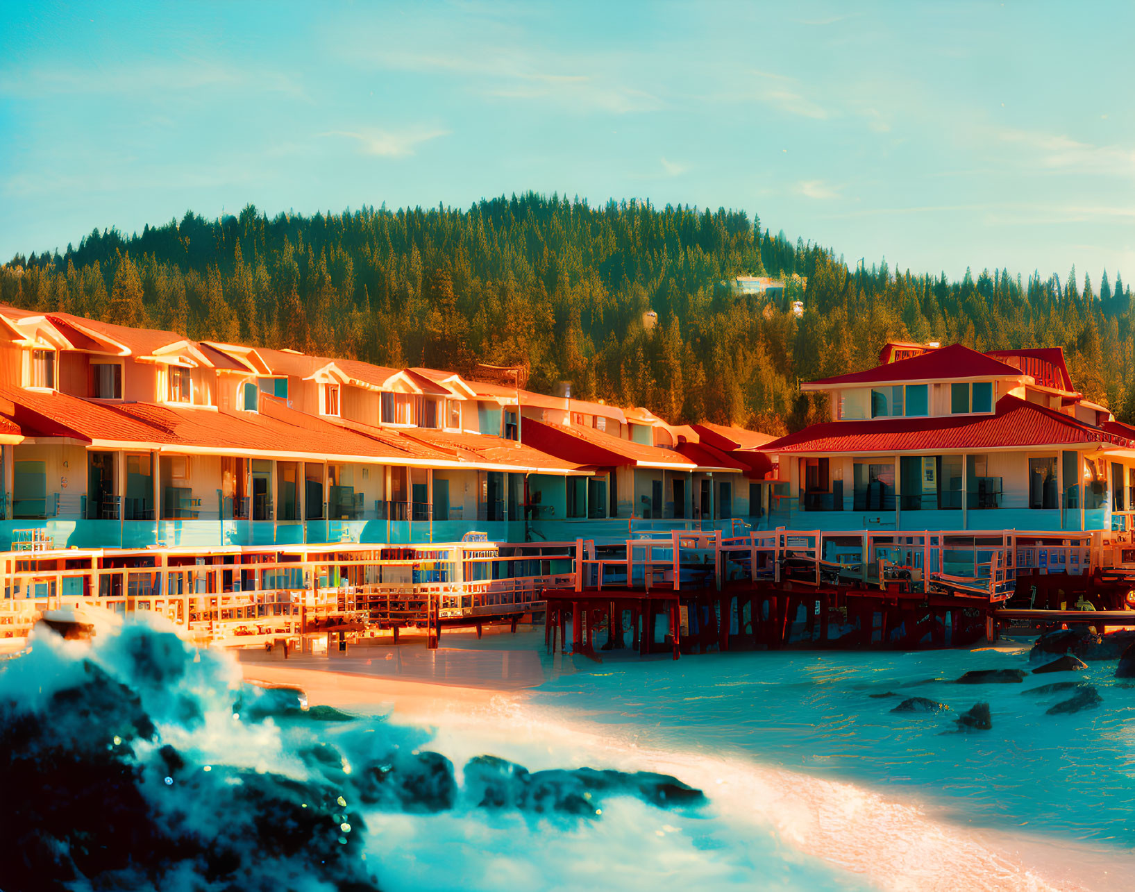 Red-roofed coastal resort on pier overlooking turquoise sea and forested hill under clear sky