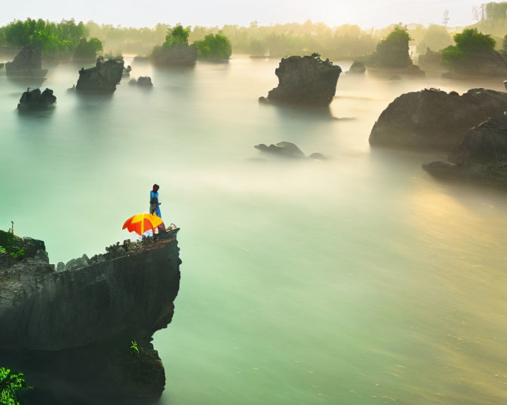 Person under red umbrella on misty river cliff with rocks and greenery.