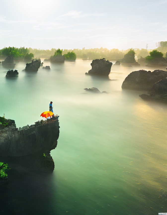 Person under red umbrella on misty river cliff with rocks and greenery.