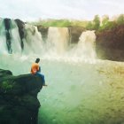 Person under red umbrella on misty river cliff with rocks and greenery.