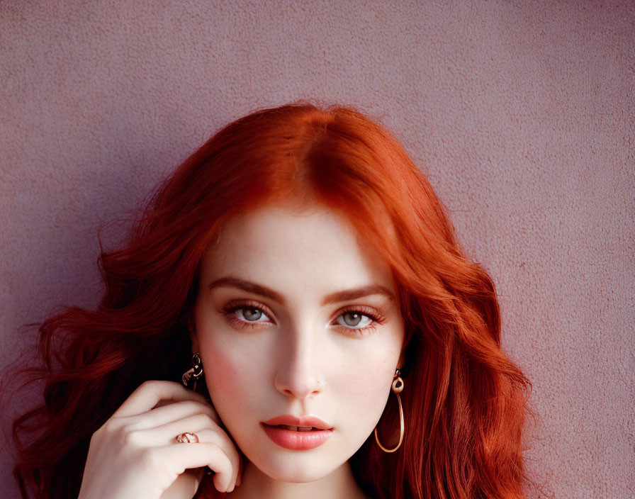 Portrait of a person with red hair, fair skin, blue eyes, and hoop earrings on pink background