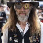 Man with long hair in hat exhales intricate smoke swirls, wearing military-style vest.