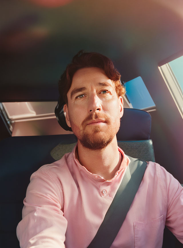 Man in pink shirt with seatbelt in car under sunlight.