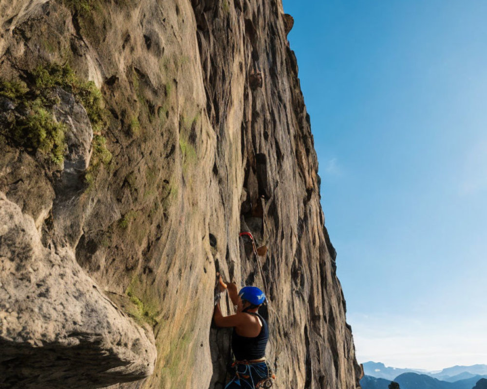 Rock climber in blue helmet ascending steep cliff against mountain backdrop