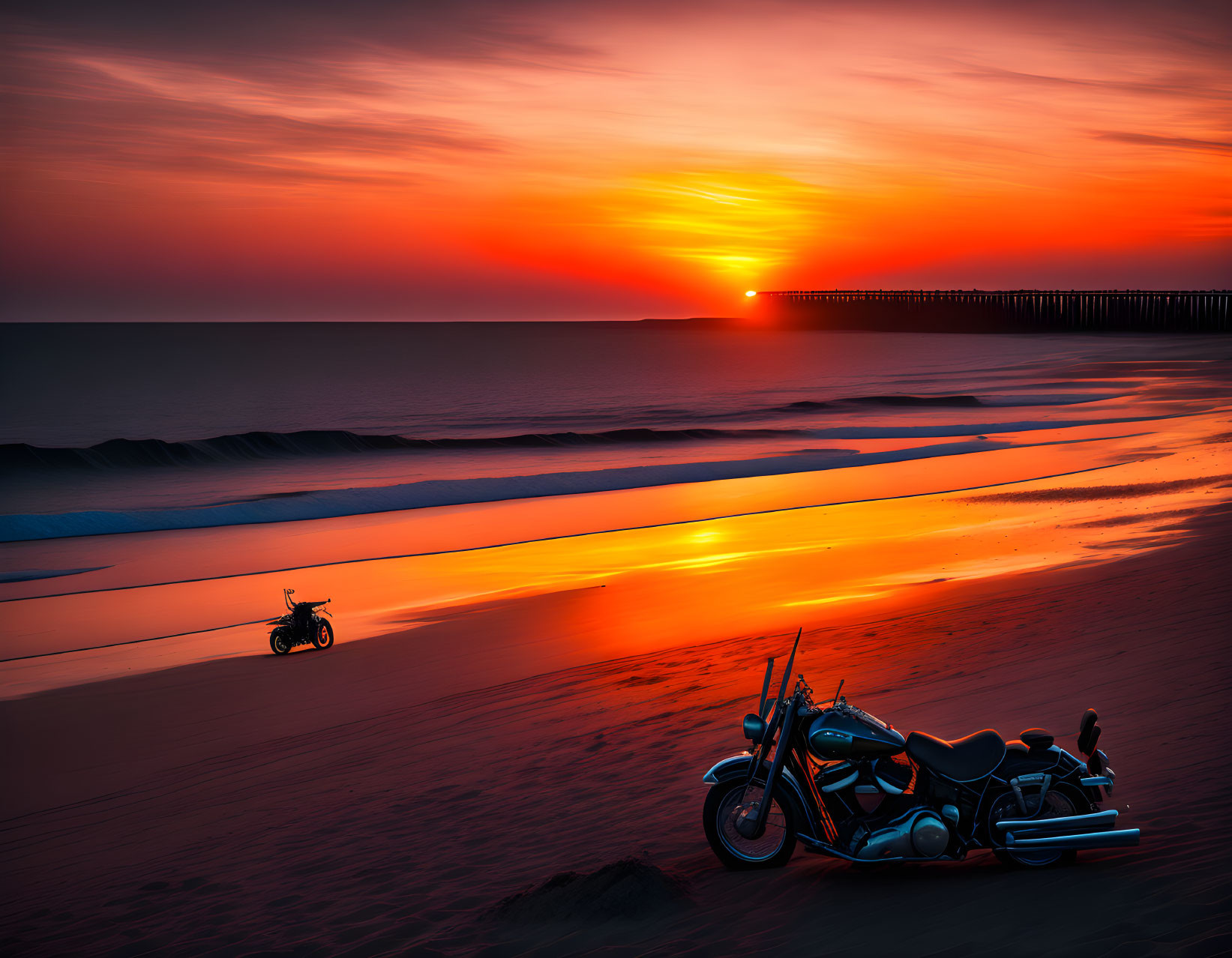 Motorcycle parked on beach at sunset with orange skies and pier.