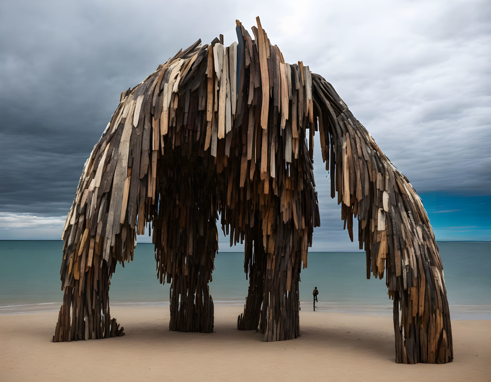 Giant wooden arch sculpture on sandy beach with lone figure under cloudy sky