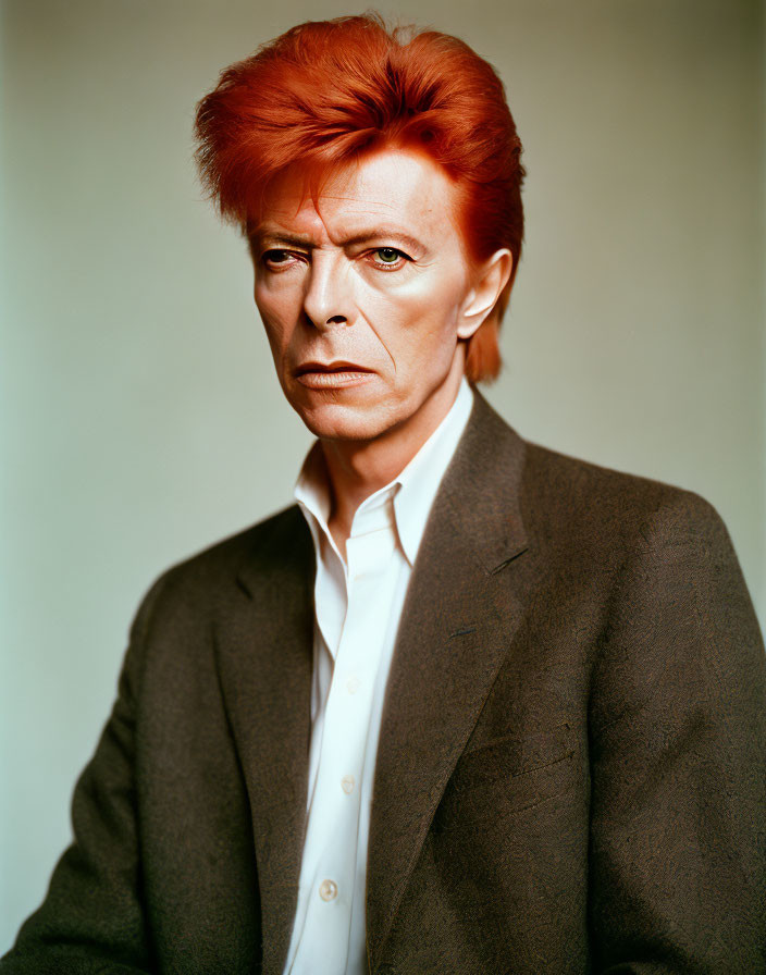 Vibrant red-haired person in white shirt and dark jacket against neutral backdrop