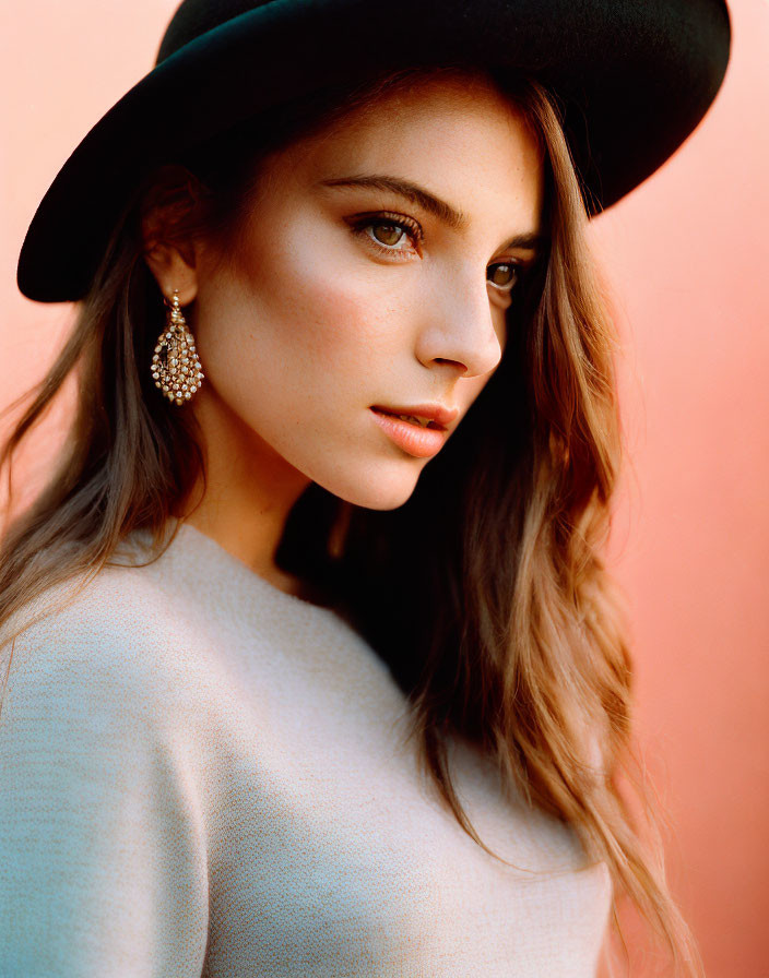 Portrait of woman with long hair in black hat and earrings against warm backdrop