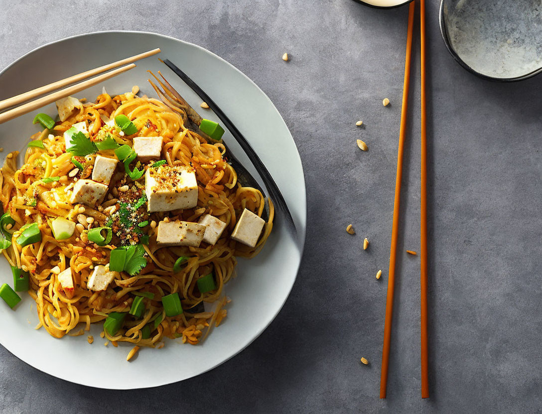 Vegan stir-fried noodles with tofu, sesame seeds, and green onions beside orange chopsticks