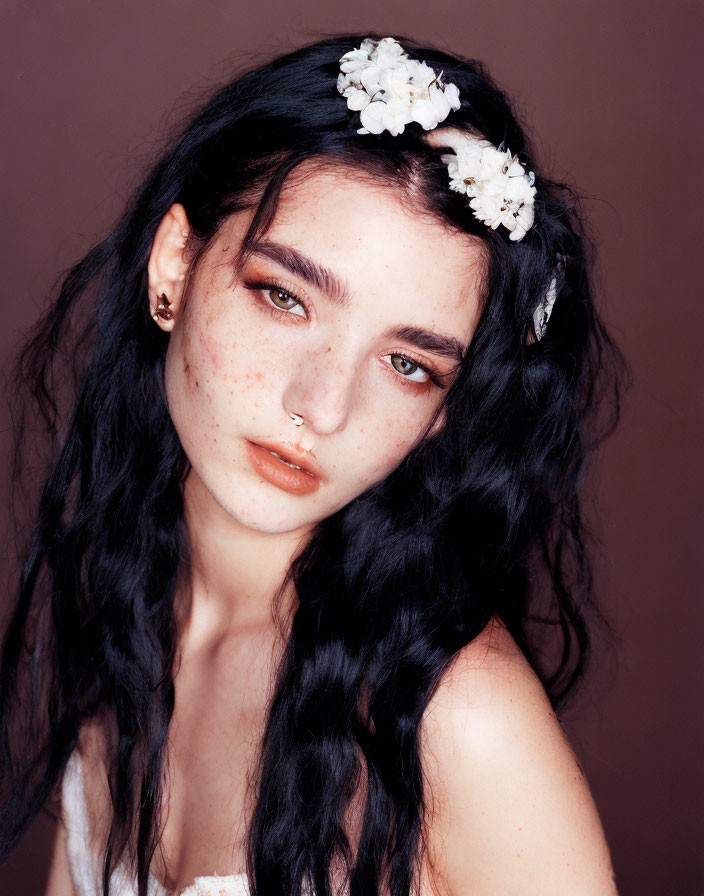 Portrait of person with freckles in white floral headband and nose ring on brown background