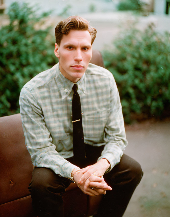 Man in Checkered Shirt and Tie Sitting on Brown Outdoor Couch with Green Shrubbery Background
