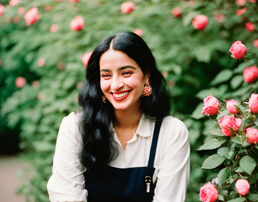 Smiling woman in white blouse and overalls by blooming rose bushes