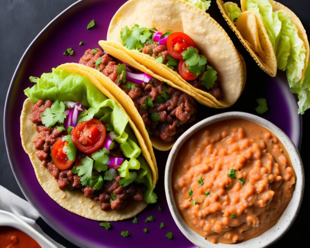 Tacos with ground meat, lettuce, tomatoes, and cilantro on purple plate with refried beans