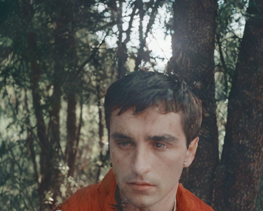 Contemplative man in red vest standing in forest with sunlight filtering through trees