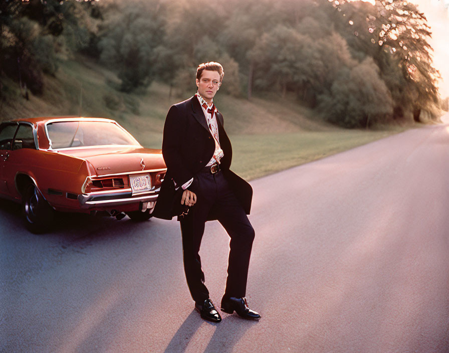 Man in suit leaning on red vintage car parked on road with trees and sunlight.