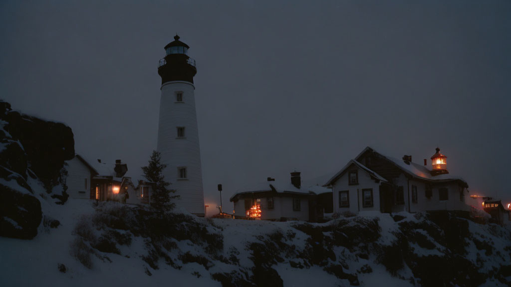 Lighthouse and buildings illuminated at twilight with snow-covered rocky terrain