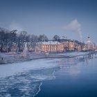 Snow-covered winter cityscape at dusk with calm river and historic buildings