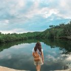 Person with backpack by tranquil river observing mirrored forest under cloudy sky