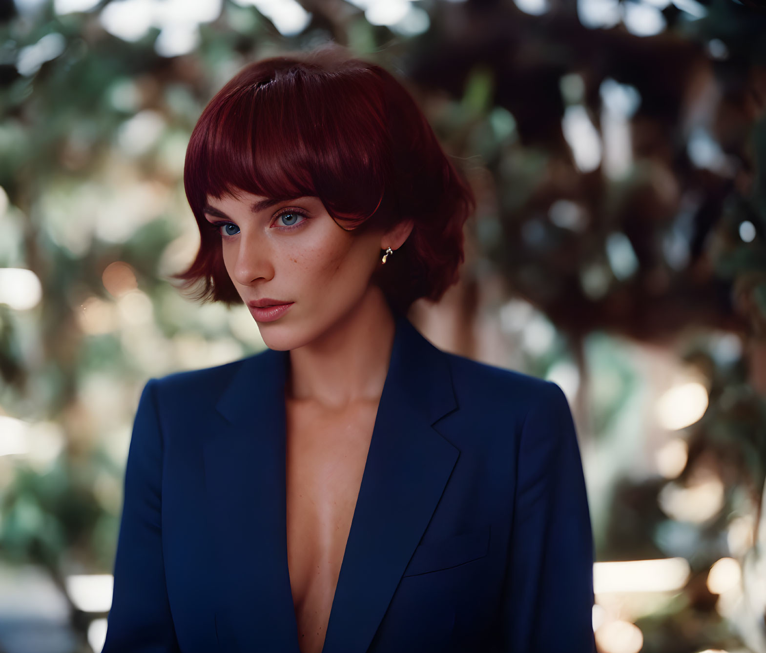 Red-haired woman in bob cut and blue blazer poses with intense gaze in dimly lit setting.