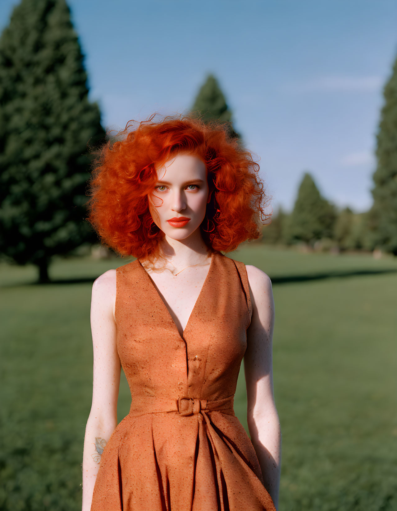 Red-haired woman in brown dress outdoors with green trees and blue sky