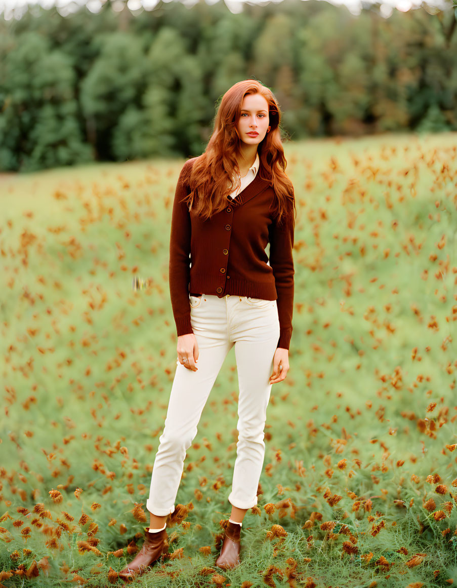 Woman in brown cardigan and white pants standing in field with tall grass and trees.