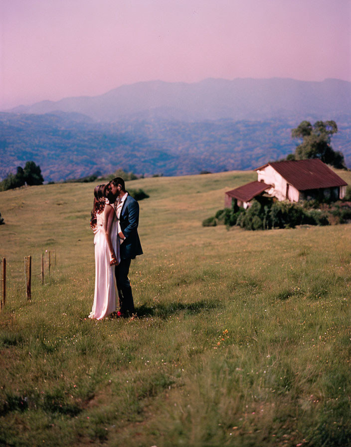 Embracing couple in green field with rolling hills and house under clear sky