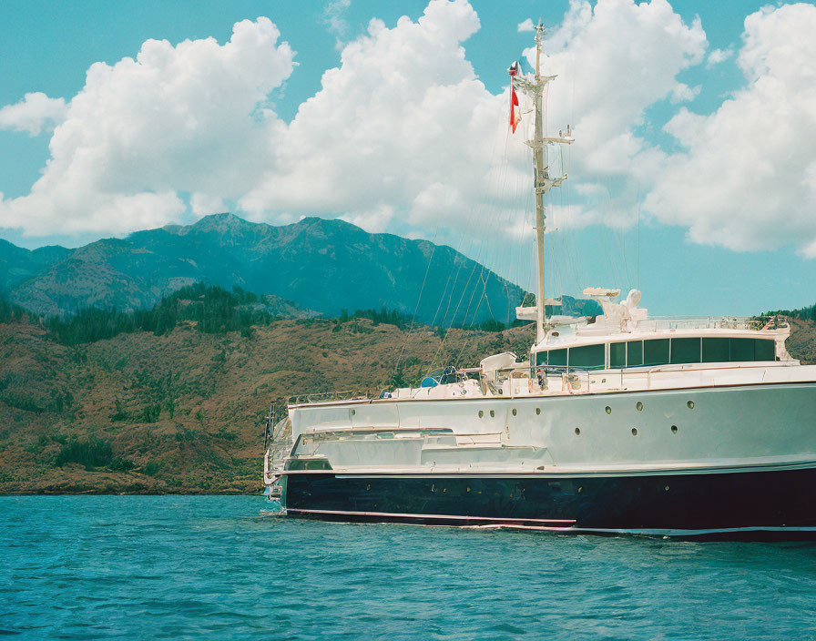 Luxury yacht near tropical coastline with mountains and fluffy clouds.